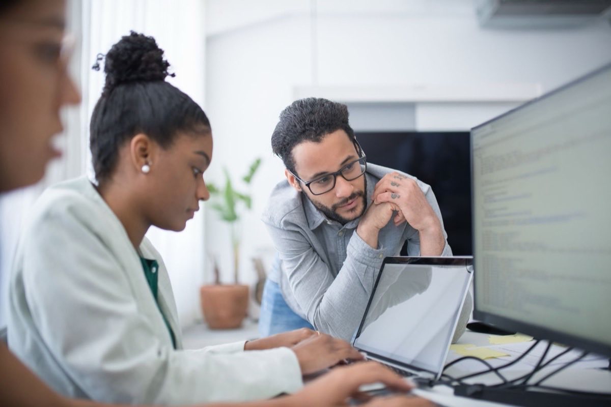 Zwei Ingenieure beugen sich gemeinsam über Laptop und Monitor, um Code in einem hellen Büro zu prüfen
