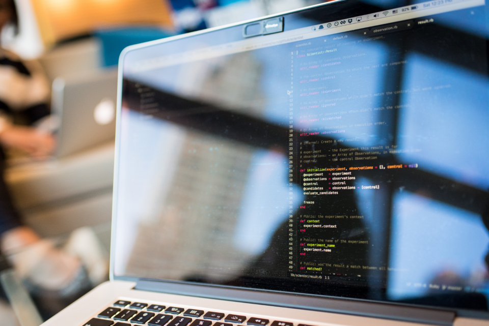 Close-up of a laptop screen showing real Ruby source code, a colleague working out of focus in the background