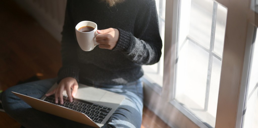 Frau am sonnigen Fenster, Kaffeetasse in der Hand, tippt auf einem Laptop auf ihrem Schoß