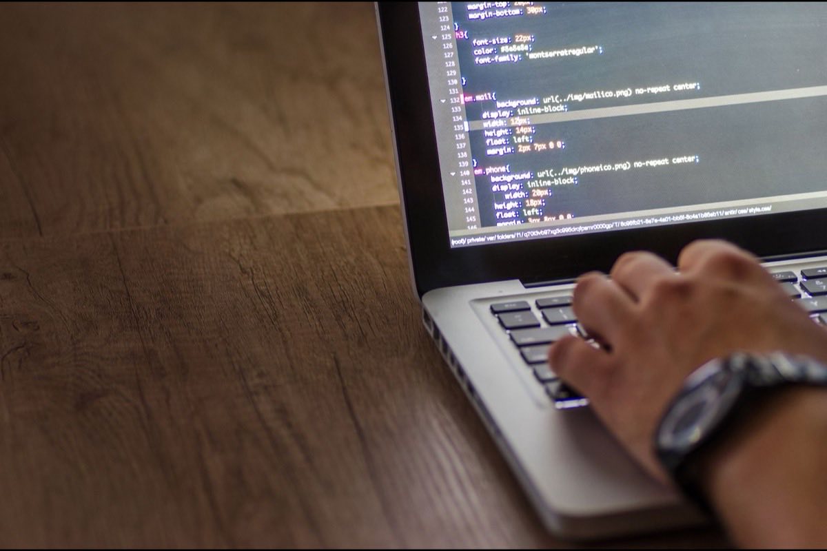 Close-up of a hand typing on a laptop displaying CSS code, on a warm wooden desk
